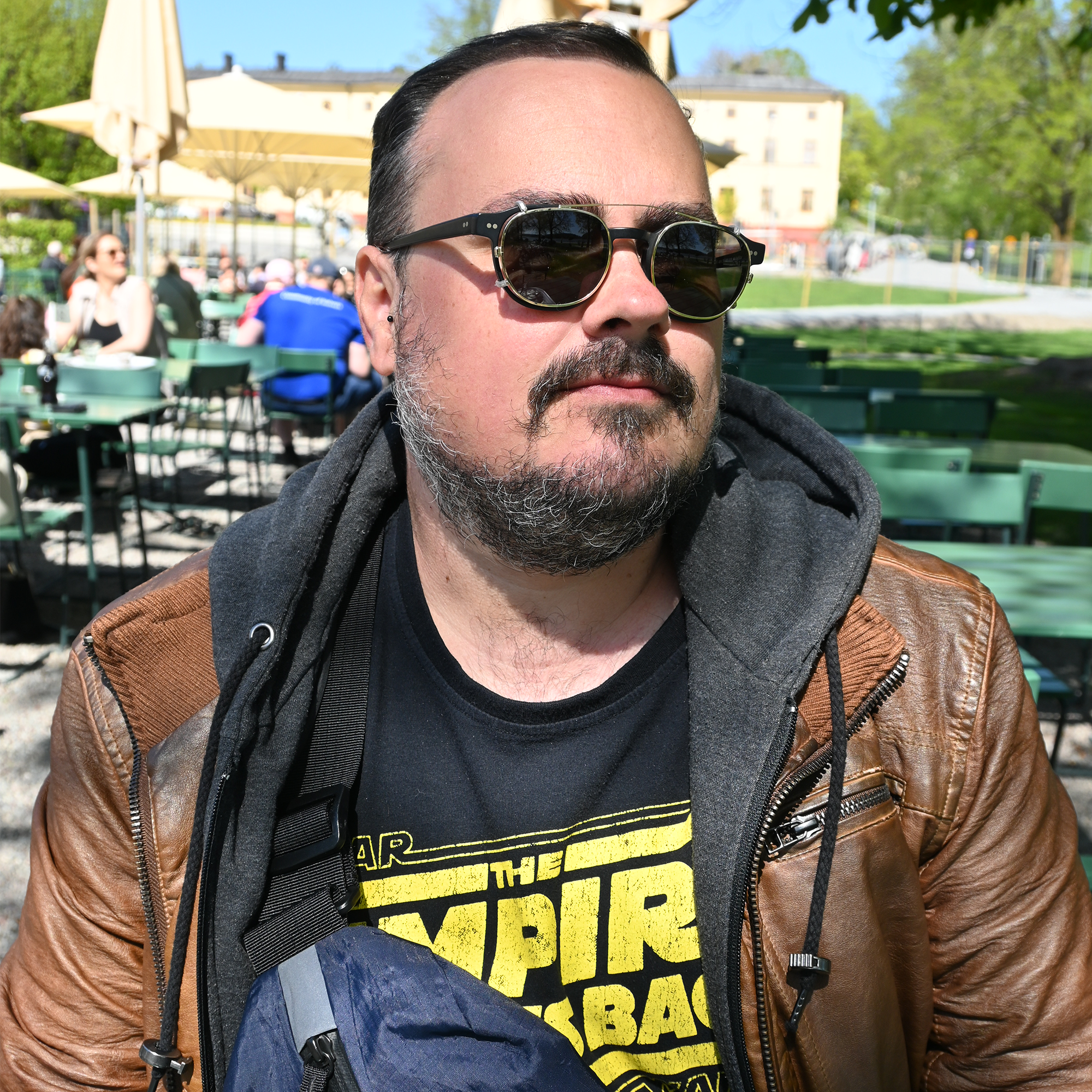 A photo of Elliott Bledsoe wearing glasses with clip on sunglasses, a black beard with a bit of grey in it, a brown leather jacket with a grey hood and a black t-shirt with the logo for Star Wars: The Empire Strikes Back on it in yellow.